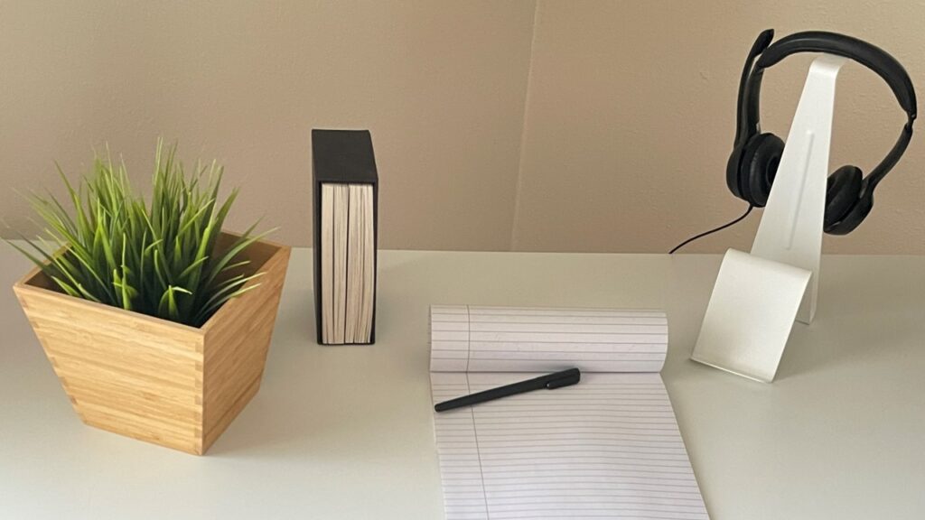 Picture of a white desk with a green plant in a wooden pot, a set of books, a notebook with a black pen and a pair of headphones resting on a headphone stand from Ikea.